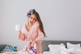Young beautiful housewife  in pink bathrobe and curler on head holding iron, speaking on phone and holding a cup. She looks astonished